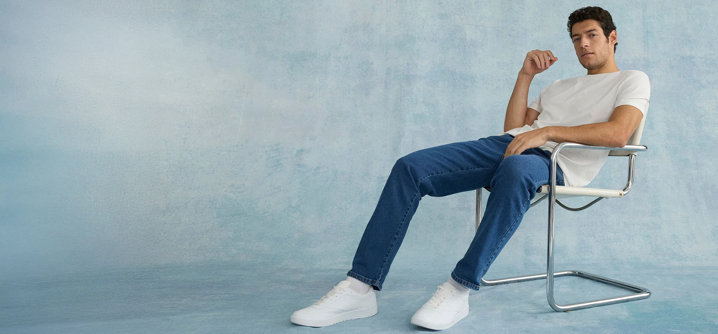 Swobodna stylizacja: biały T-shirt, niebieskie jeansy i białe sneakersy na nowoczesnym krześle.