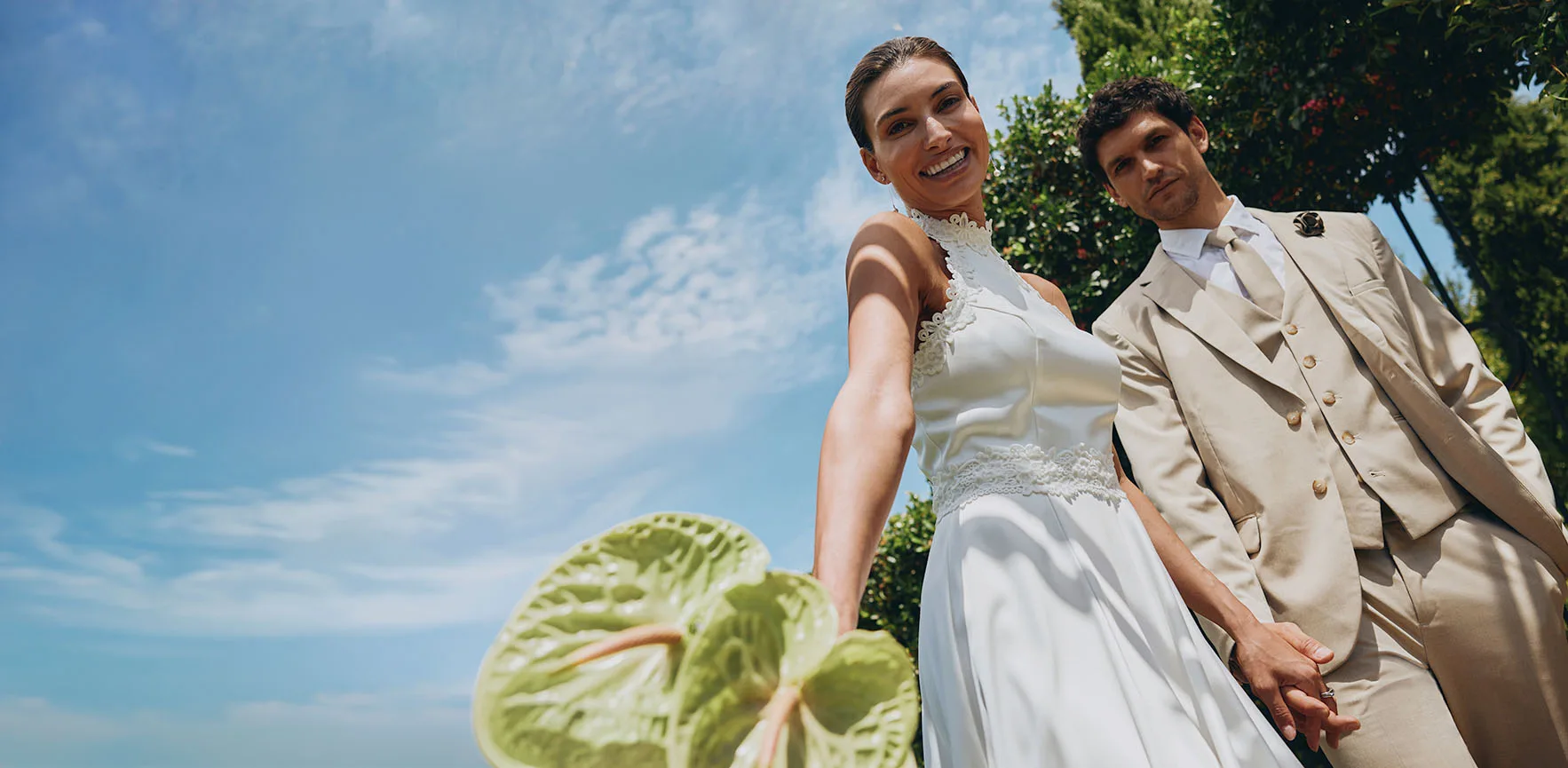 Para młoda skąpana w ciepłych promieniach słońca: ona w klasycznej białej sukni, on w beżowym garniturze i stylowych okularach przeciwsłonecznych.