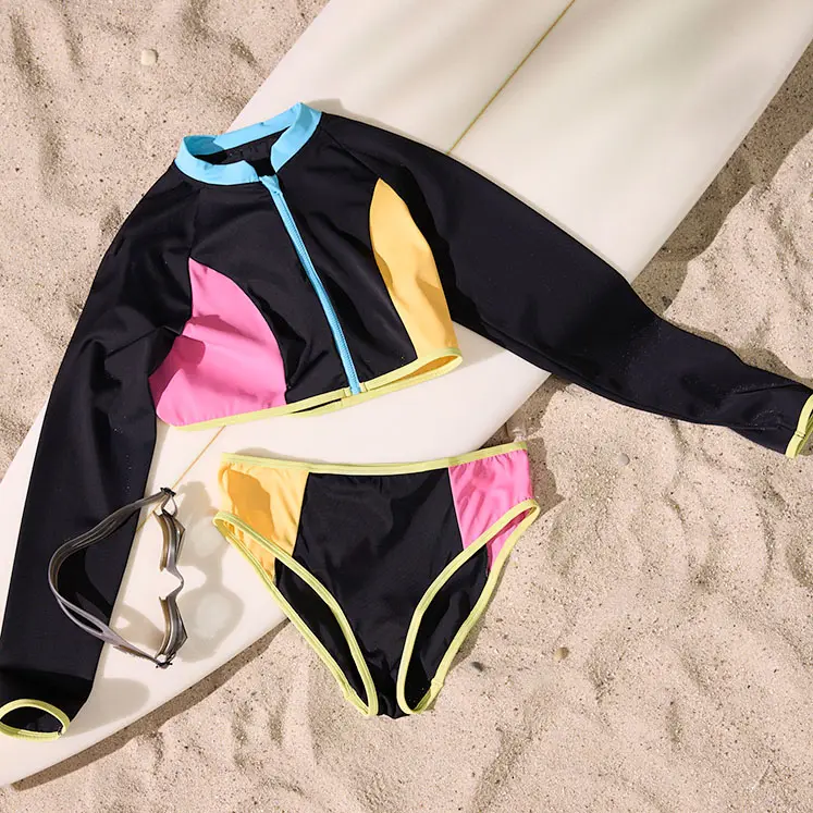 Kolorowy strój kąpielowy z długim rękawem i bikini na piasku obok deski surfingowej.