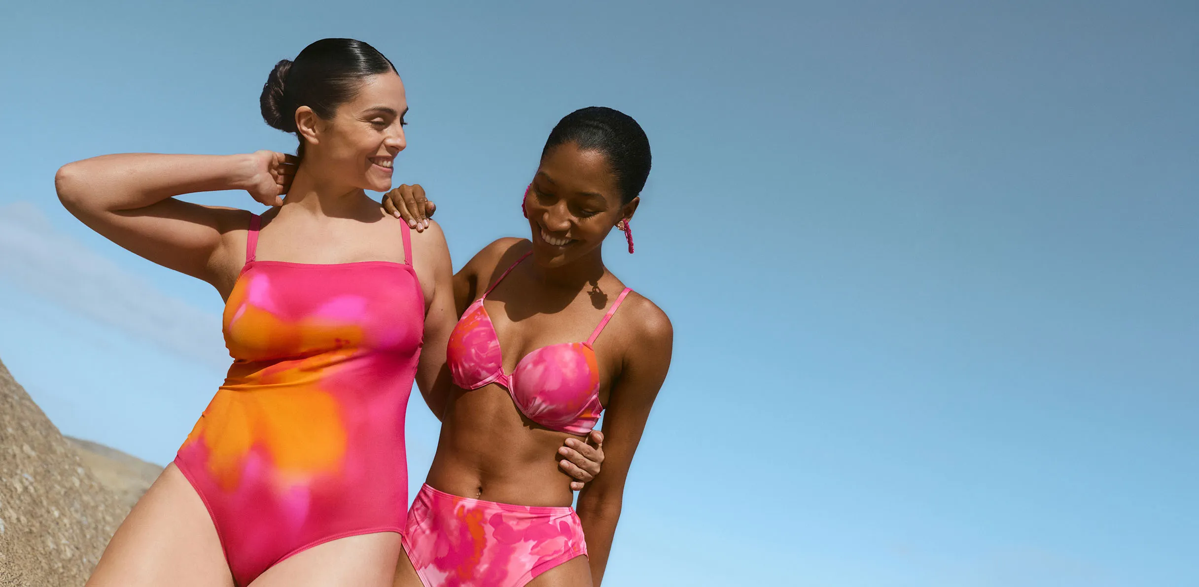 Dwie kobiety na plaży ubrane w odblaskowe, różowe stroje kąpielowe i bikini.